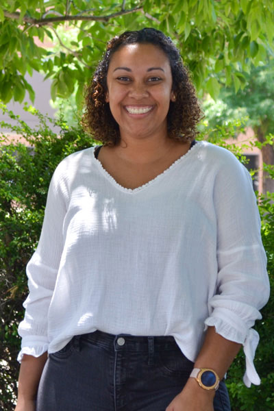 A woman smiling at the camera, standing outdoors with trees behind her, wearing a white blouse and black pants.