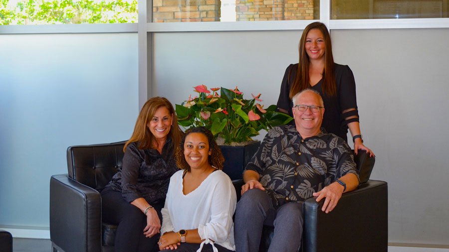 The image shows a group of four people posing together in an office setting, with a woman seated on the left, a man standing next to her, another man seated on the right, and a woman standing between them. They are all dressed in semi-formal attire and appear to be in a professional environment.