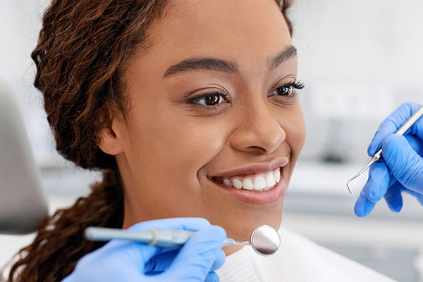 The image depicts a smiling person wearing a white lab coat and a stethoscope around their neck, holding a dental mirror with a dental pick, likely in a dental office setting.