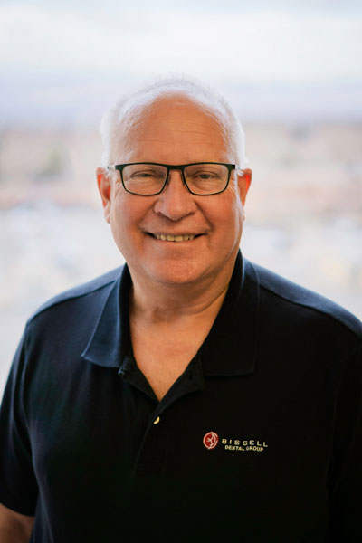 The image shows a man wearing glasses, smiling slightly, with gray hair, dressed in a dark shirt with a logo on the left side of his chest, standing against a blurred background that appears to be an indoor setting with artificial lighting.