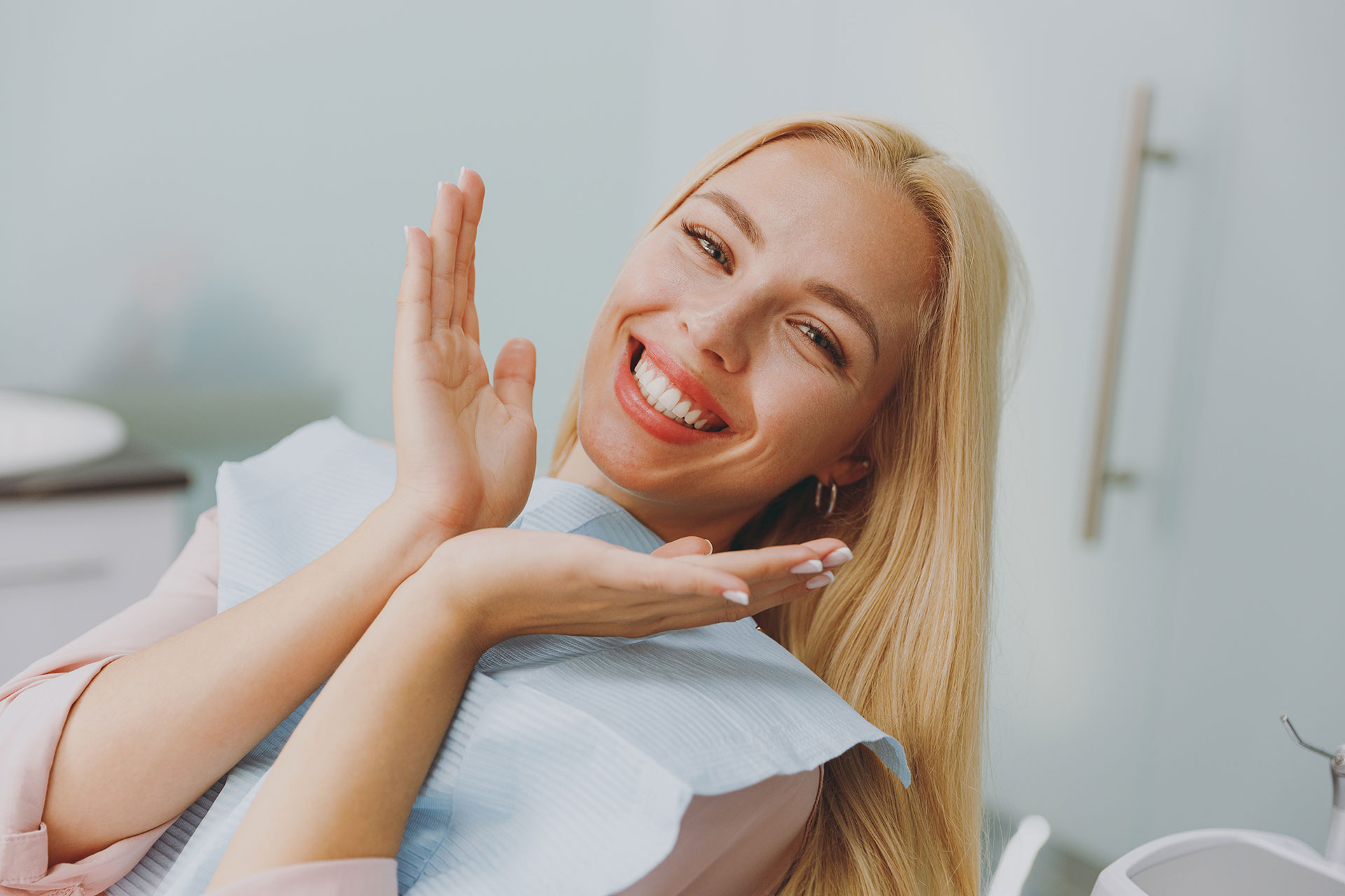 The image shows a woman with blonde hair sitting in a dental chair, smiling broadly, with her hands clasped together near her chest.