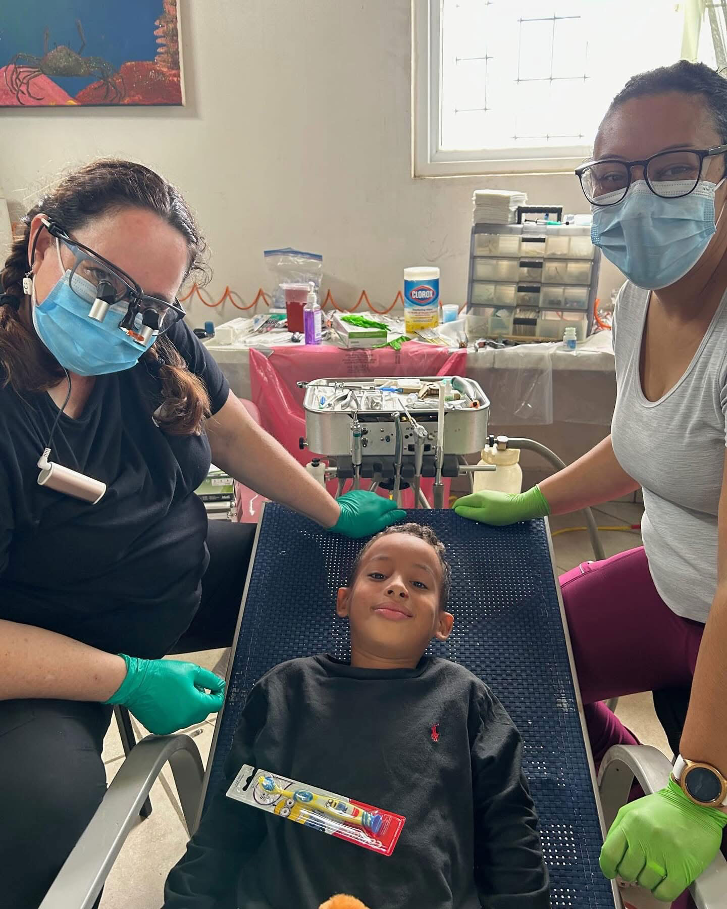 In the image, three individuals are wearing protective face masks and gloves while standing around a table with medical equipment. One person is seated on a chair with a blue backrest, looking up at the camera, while another person is holding up a child's head for examination. The setting appears to be an indoor medical or dental facility.
