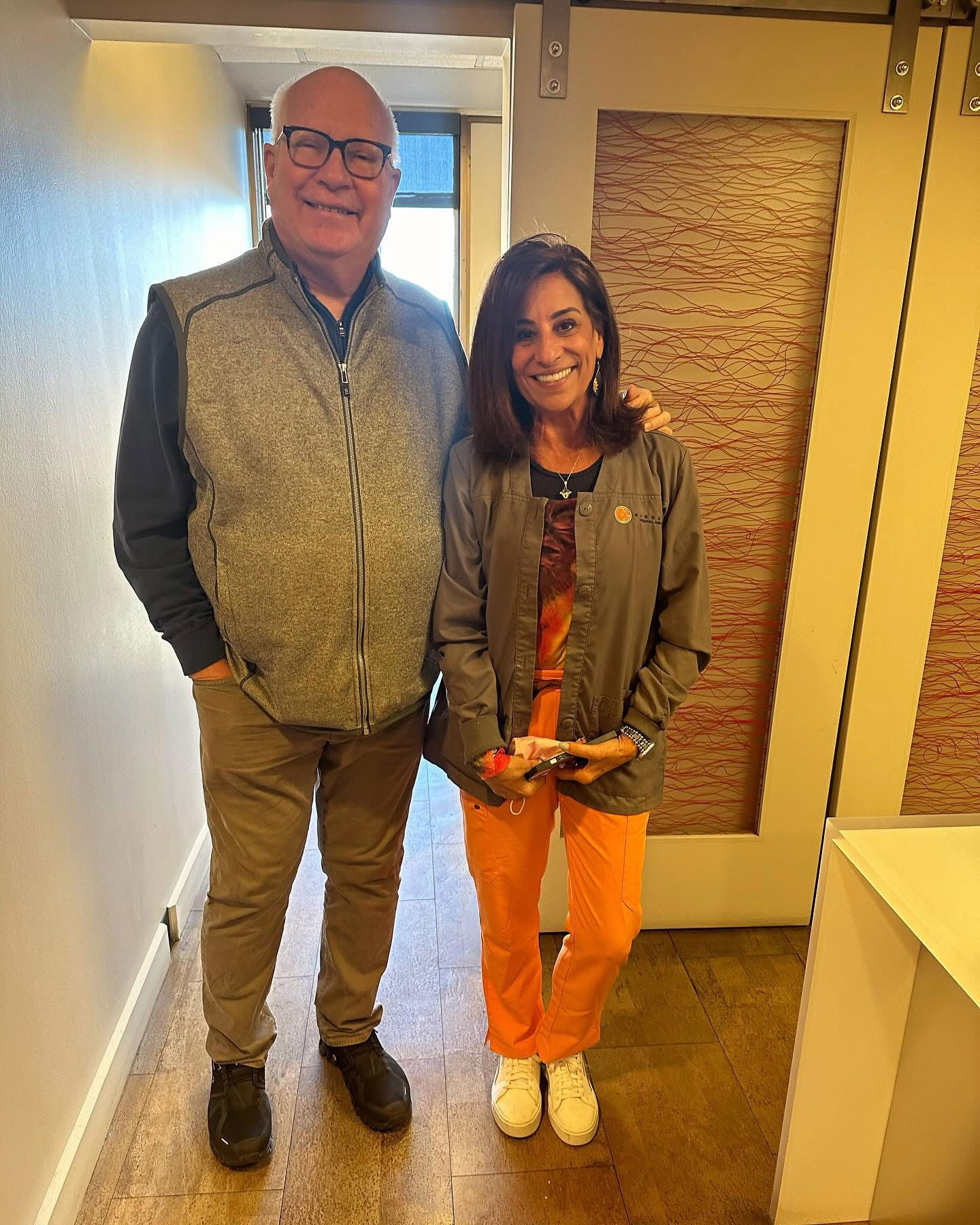 A man and woman standing together indoors, posing for a photo with smiles on their faces  the man has a beard and wears glasses, while the woman has short hair and is dressed in casual attire with an orange top and blue jeans.