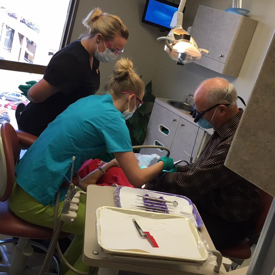In this image, there are four individuals present  three women wearing surgical masks and engaged in dental procedures, and an older man with a white beard sitting in a dentist s chair, receiving dental care.