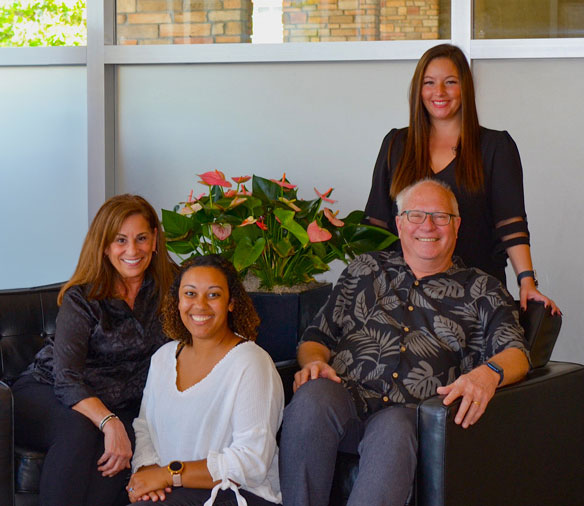 The image features a group of four individuals posing together, with three seated on a couch and one standing behind them, all smiling and looking towards the camera. They are in an indoor setting that appears to be a professional environment, likely an office or lobby, as suggested by the modern furniture and decor.