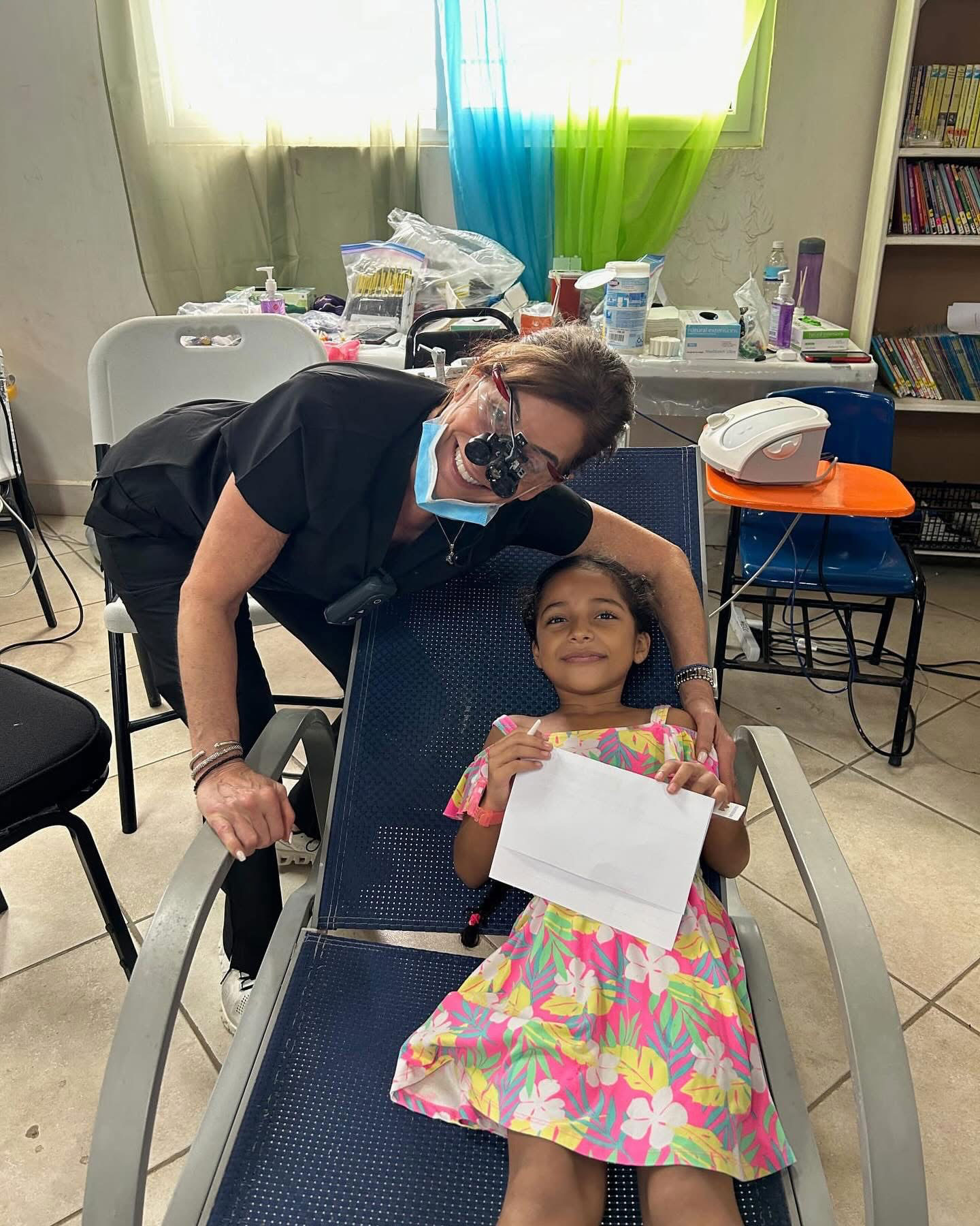 Woman sitting on blue chair with child on her lap, both wearing face masks; woman holding paper.