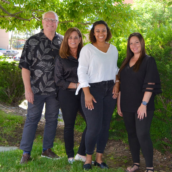 The image shows a group of four people posing together outdoors. From left to right, there is an older man wearing glasses, a woman, a young adult woman, and another young adult woman. They are standing in front of a tree with green leaves, and the background includes a building and a clear sky.