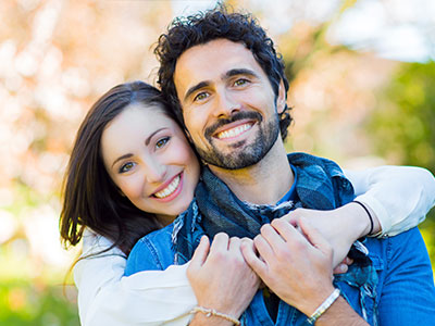 The image shows a man and woman embracing each other with smiles, posing for a photograph outdoors during daylight.