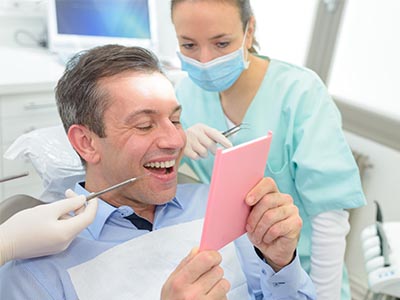 The image shows a man sitting in a dental chair with a big smile on his face, holding up a pink card with both hands while looking at it, surrounded by dental professionals who appear engaged and attentive to him.