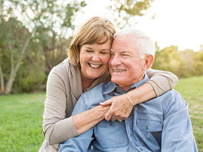 The image shows an elderly couple embracing each other outdoors during daylight, with both individuals smiling.