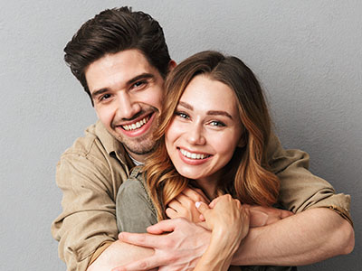 A man and woman are embracing each other in a warm pose, smiling at the camera, with the man wearing a button-up shirt and the woman in a casual top.