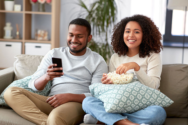 A man and woman sitting on a couch, smiling and enjoying popcorn while watching something on their phones.