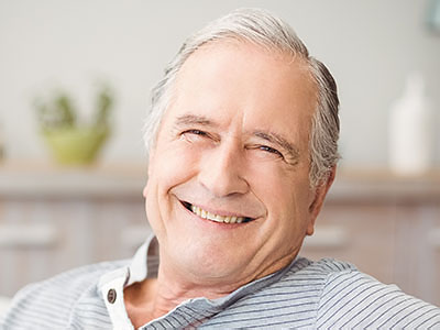 The image shows an elderly man with white hair, smiling broadly at the camera, wearing a light blue shirt, sitting comfortably in a home setting with a warm ambiance.