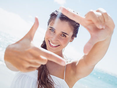 The image features a smiling woman with long hair, holding up her index finger as if taking a photo, against a bright, sunny background.